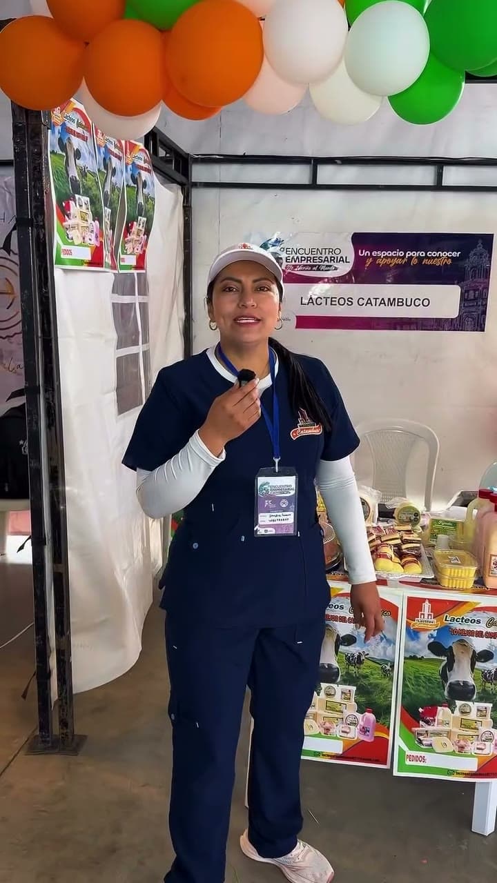 Vocera de Lácteos Catambuco con uniforme azul y gorra frente al stand de la feria, bajo un arco de globos naranjas, blancos y verdes y un aviso con el nombre de la marca